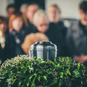 A metal urn with ashes of a dead person on a funeral, with people mourning in the background on a memorial service. Sad grieving moment at the end of a life. Last farewell to a person in an urn.